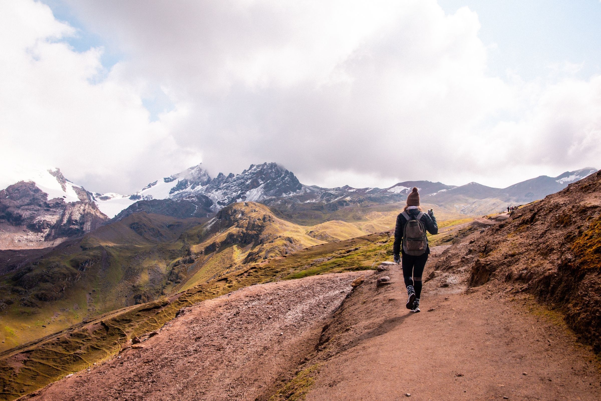Viajera de espaldas caminando por un sendero de los Andes de Cusco, con nevados al fondo